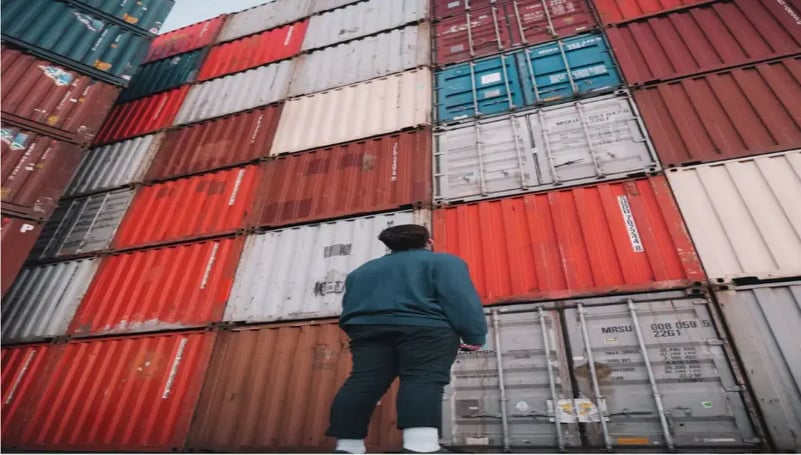 man-in-black-jacket-standing-in-front-of-red-and-blue-intermodal-containers