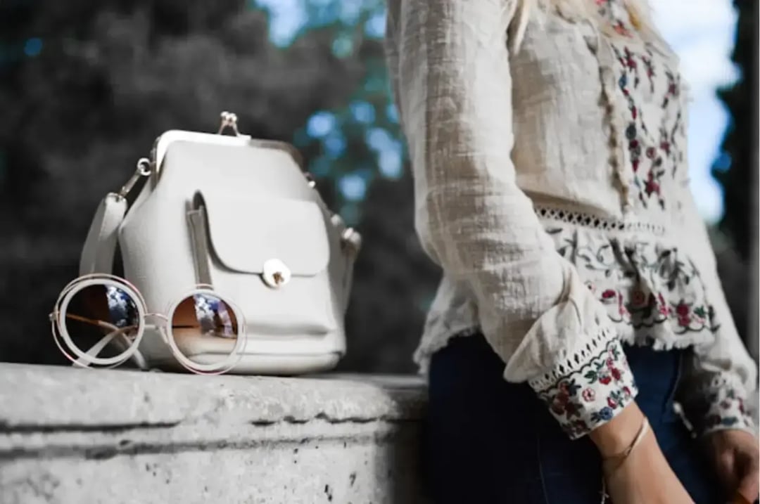 woman-wearing-beige-and-red-floral-top-leaning-on-gray-concrete-slab-with-white-leather-bag-ontop